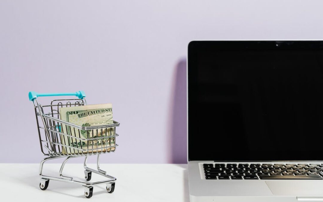 A tiny shopping cart with money bills in it standing next to a laptop