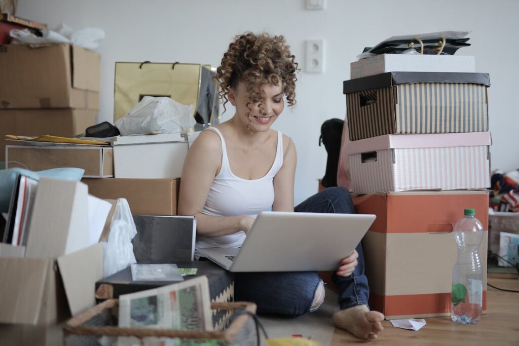 Woman sitting on the floor surrounded by boxes and clutter and researching how to make money by reselling your old furniture and appliances on her laptop