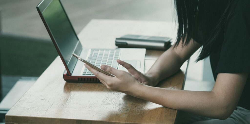 A woman looking at her phone next to a laptop.